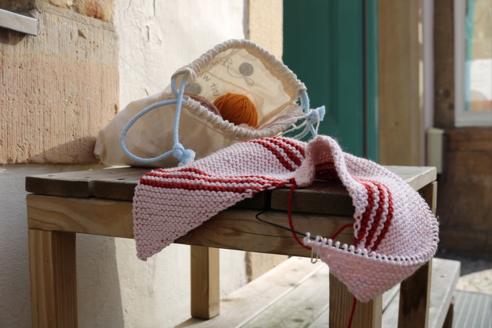 An unfinished knitted shawl still on the knitting needles lies on a coffee table together with a project bag.